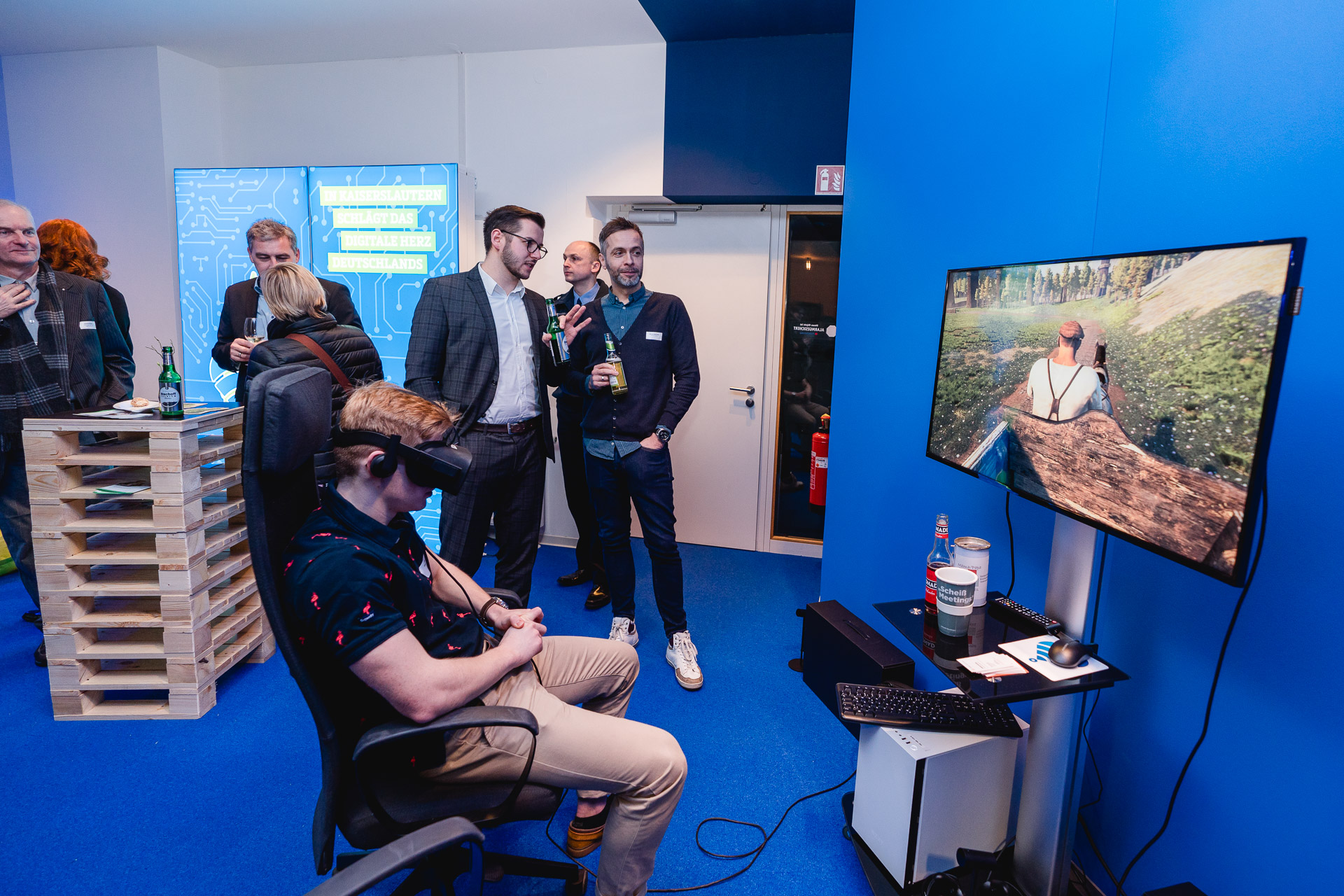 Ein junger Mann sitzt mit einer VR-Brille auf dem Kopf auf einem Stuhl. Auf einem Monitor sieht man die Rückenansicht eines Kutschers.