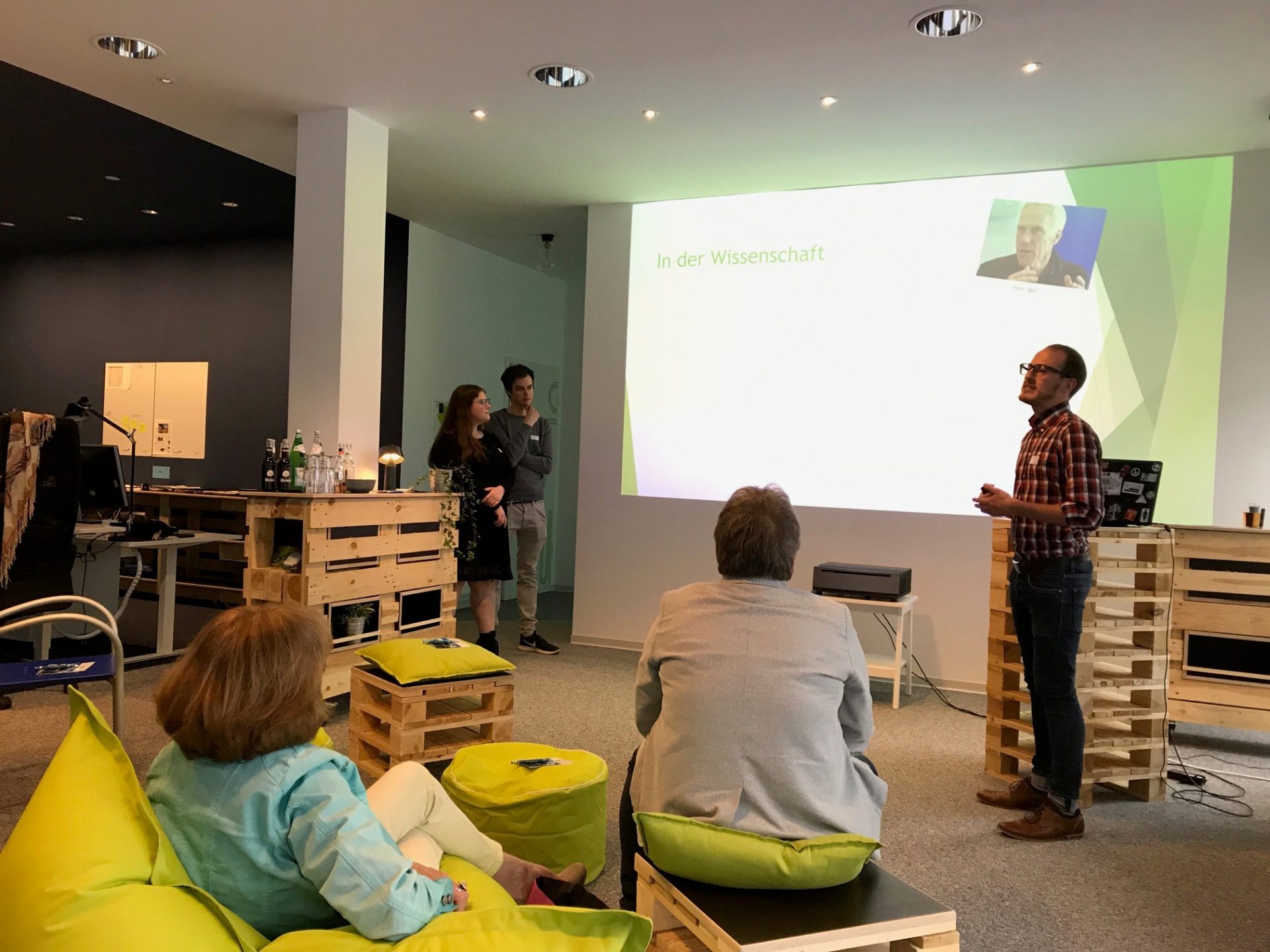 Ein Mann steht vor einem kleinen Publikum und hält einen Vortrag. Auf der Präsentation im Hintergrund steht "In der Wissenschaft". Zwei weitere Vortragende stehen am Rand.