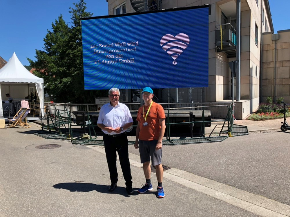 Mitarbeiter Martin Wilke steht zusammen mit Innenminister Roger Lewentz vor einer Präsentationsleinwand auf dem Festgelände in Annweiler. Auf der Leinwand steht "Die Social Wall wird Ihnen präsentiert von der KL.digital GmbH"