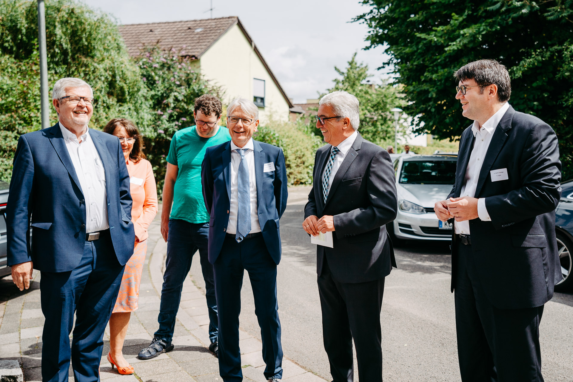 Vier Männer stehen im Bildvordergrund auf einem Gehweg und sind fröhlich.