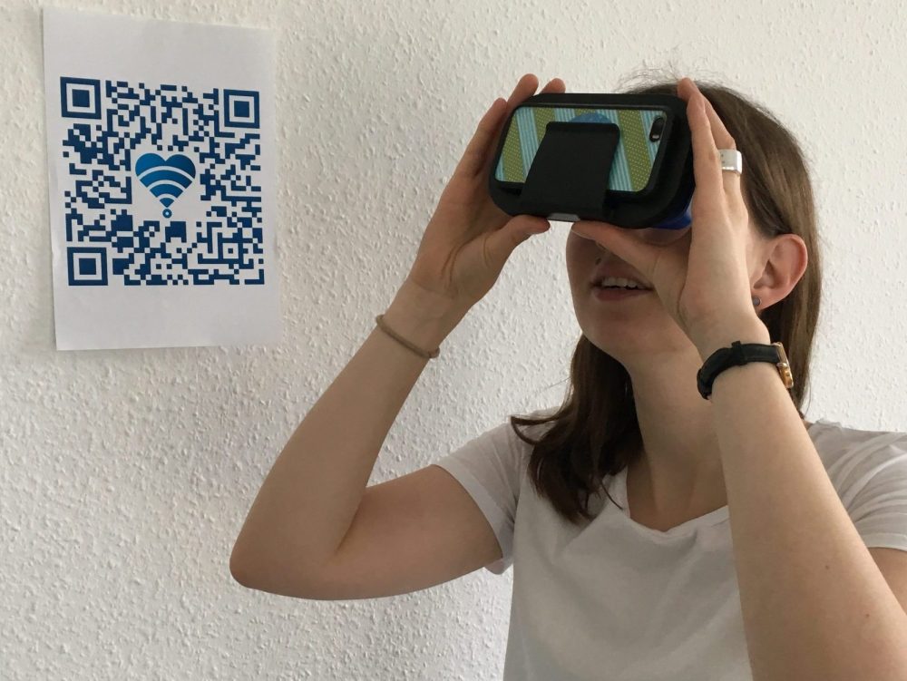 Eine Frau steht vor einem ausgedruckten QR-Code und schaut durch ein Cardboard