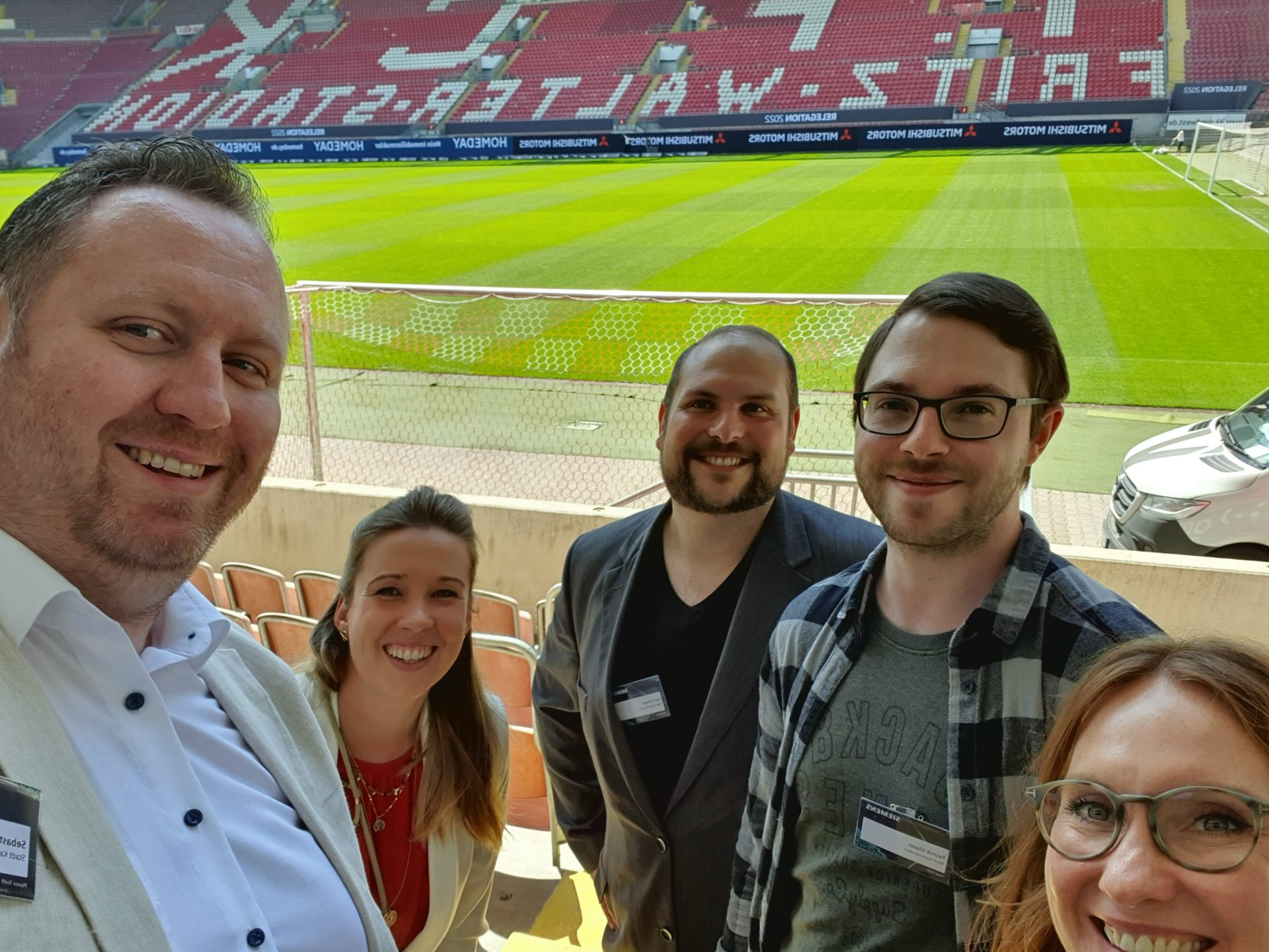 Drei Männer und zwei Frauen stehen auf der Tribüne vor einem leeren Fußballspielfeld und lächeln in die Kamera