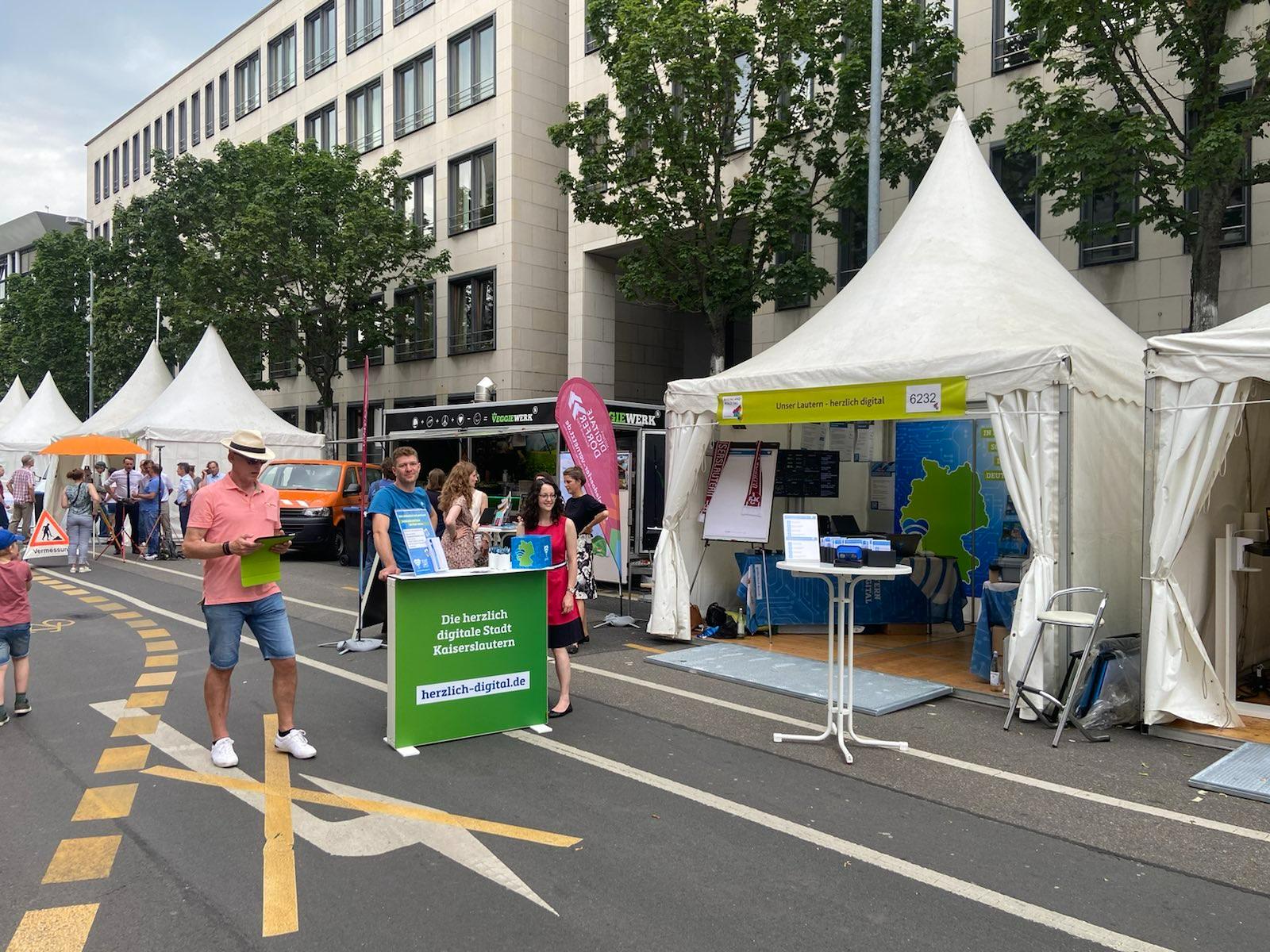 Mehrere Personen stehen auf einer Straße bei einem Fest (Rheinland-Pfalz-Tag), neben ihnen sind geöffnete Pagodenzelte zu sehen.