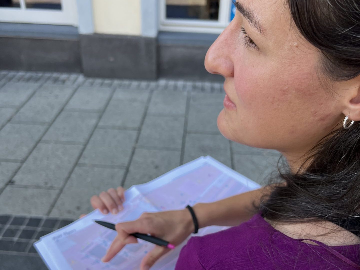 Eine Frau steht auf einem Platz und schaut in die Ferne. In den Händen hat sie einen Lageplan und einen Stift.