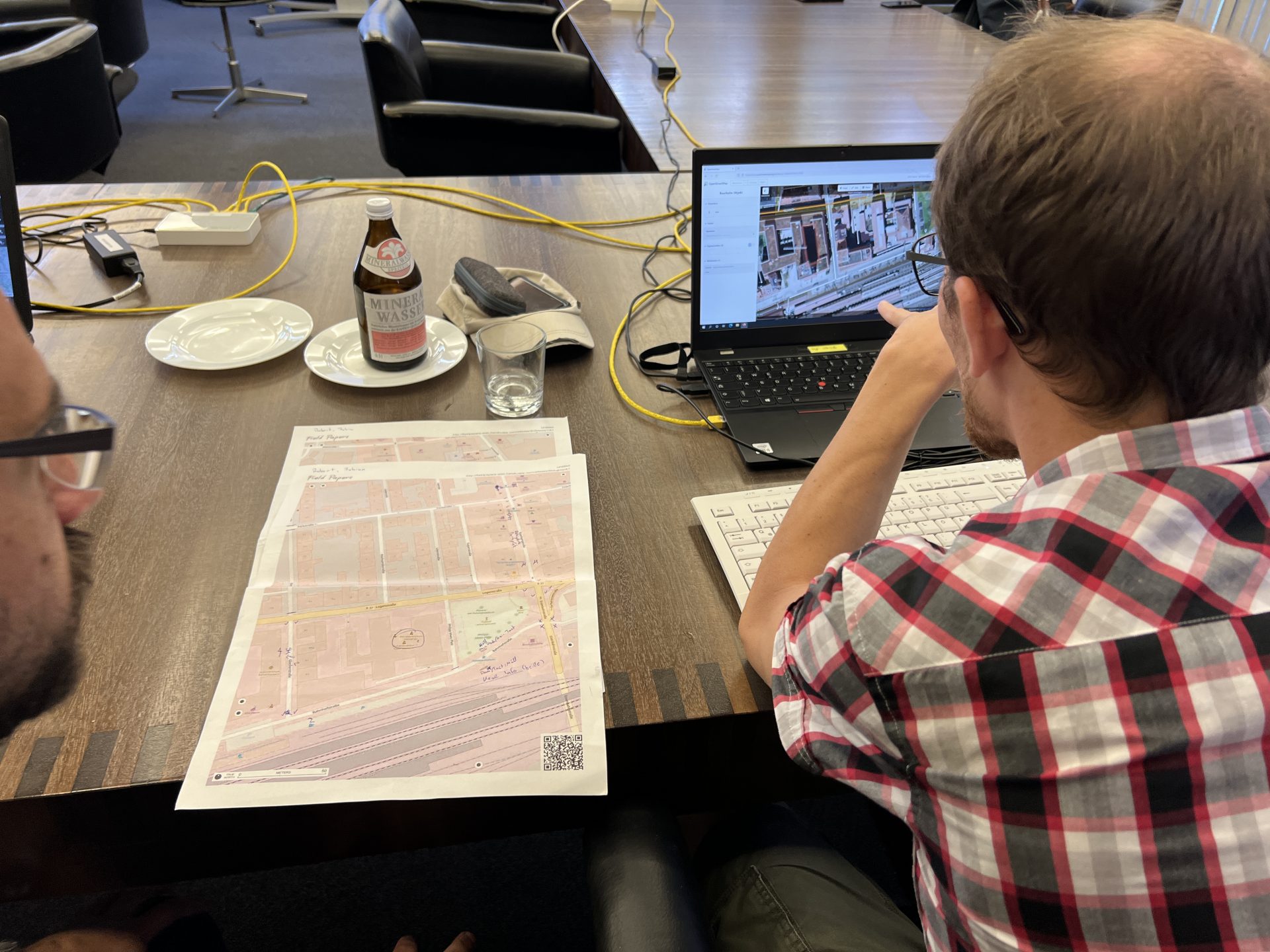 Zwei Männer betrachten eine Straßenansicht auf einem Laptop. Neben ihnen liegen Stadtpläne.