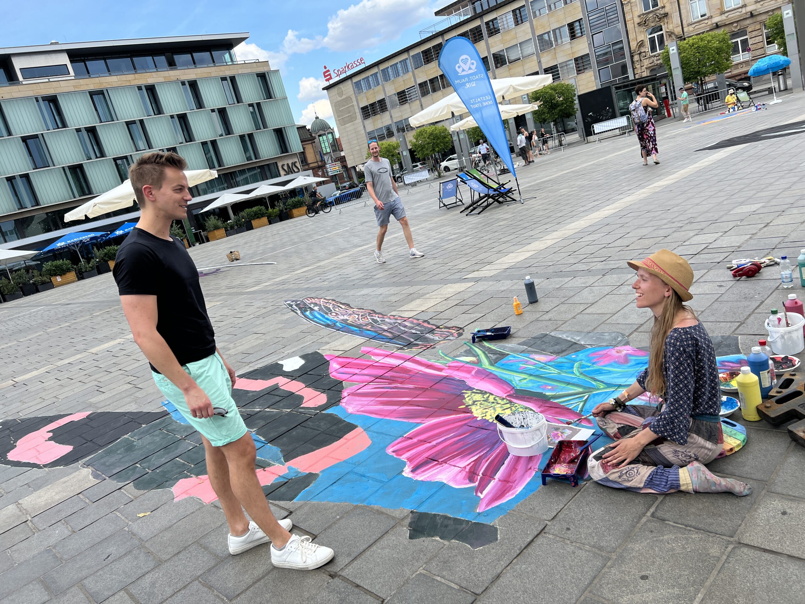 Ein Mann und eine Frau unterhalten sich. Sie sitzt vor einem Straßenmalereibild auf dem Boden.