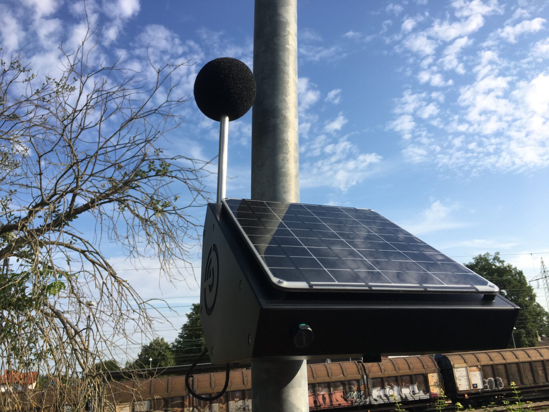 Ein kleines Solarpanel an einem Laternenmast, im Hintergrund ein Güterzug und blauer Himmel