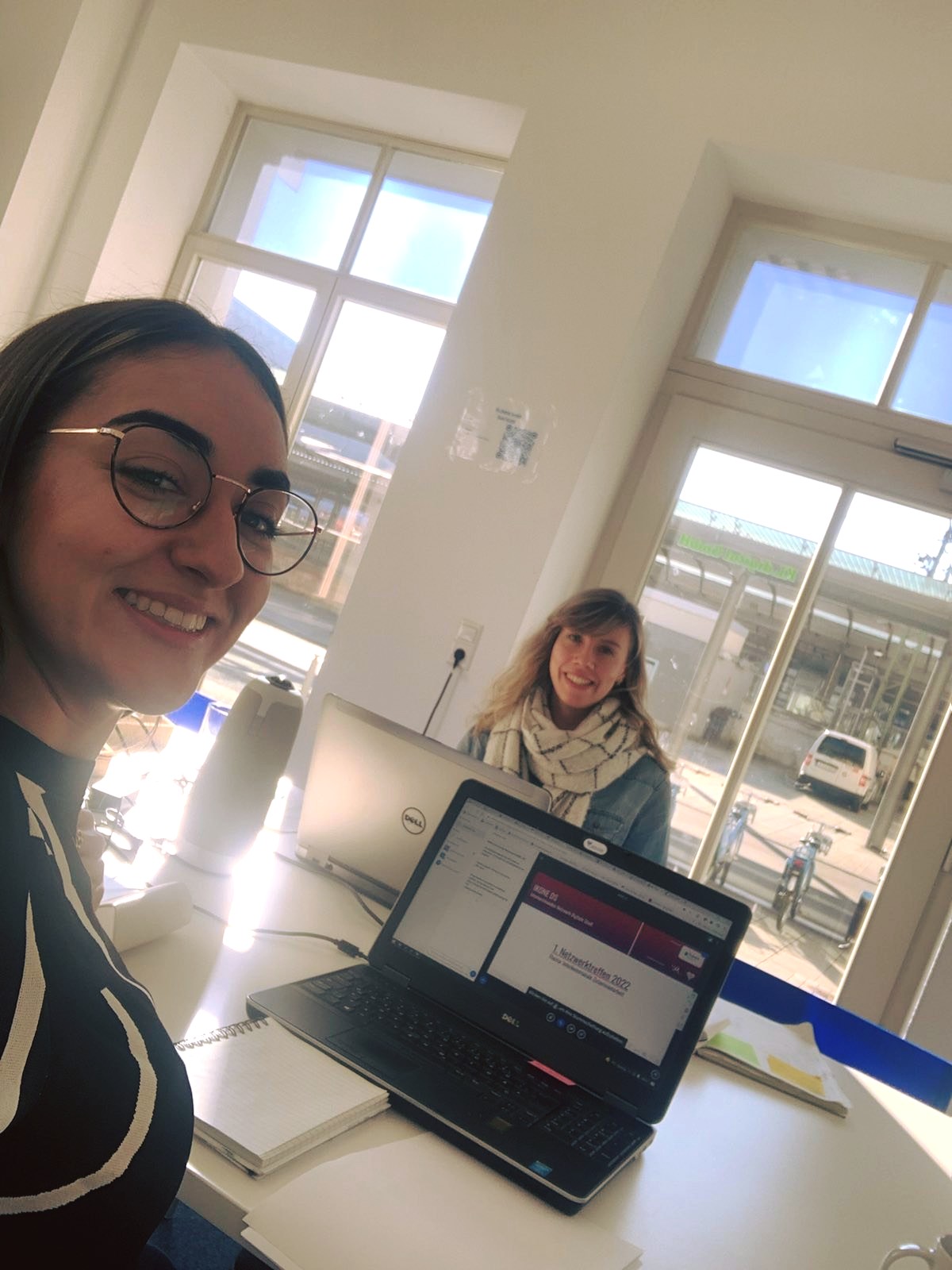 Zwei Frauen sitzen vor aufgeklappten Laptops gegenüber an einem Tisch. Sie schauen in die Kamera.