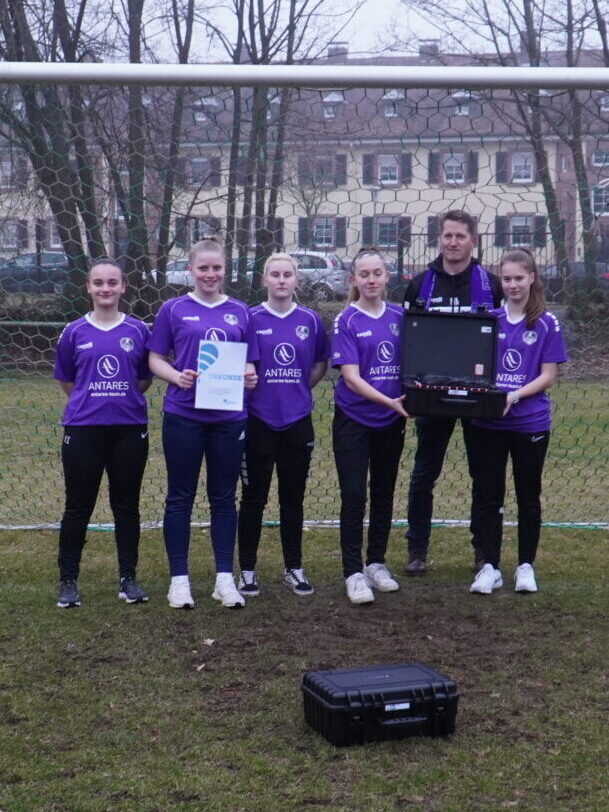 Fünf Mädchen in Triktos stehen gemeinsam mit einem Mann in einem Fußballtor auf einem Fußballplatz und halten eine Urkunde in der Hand.