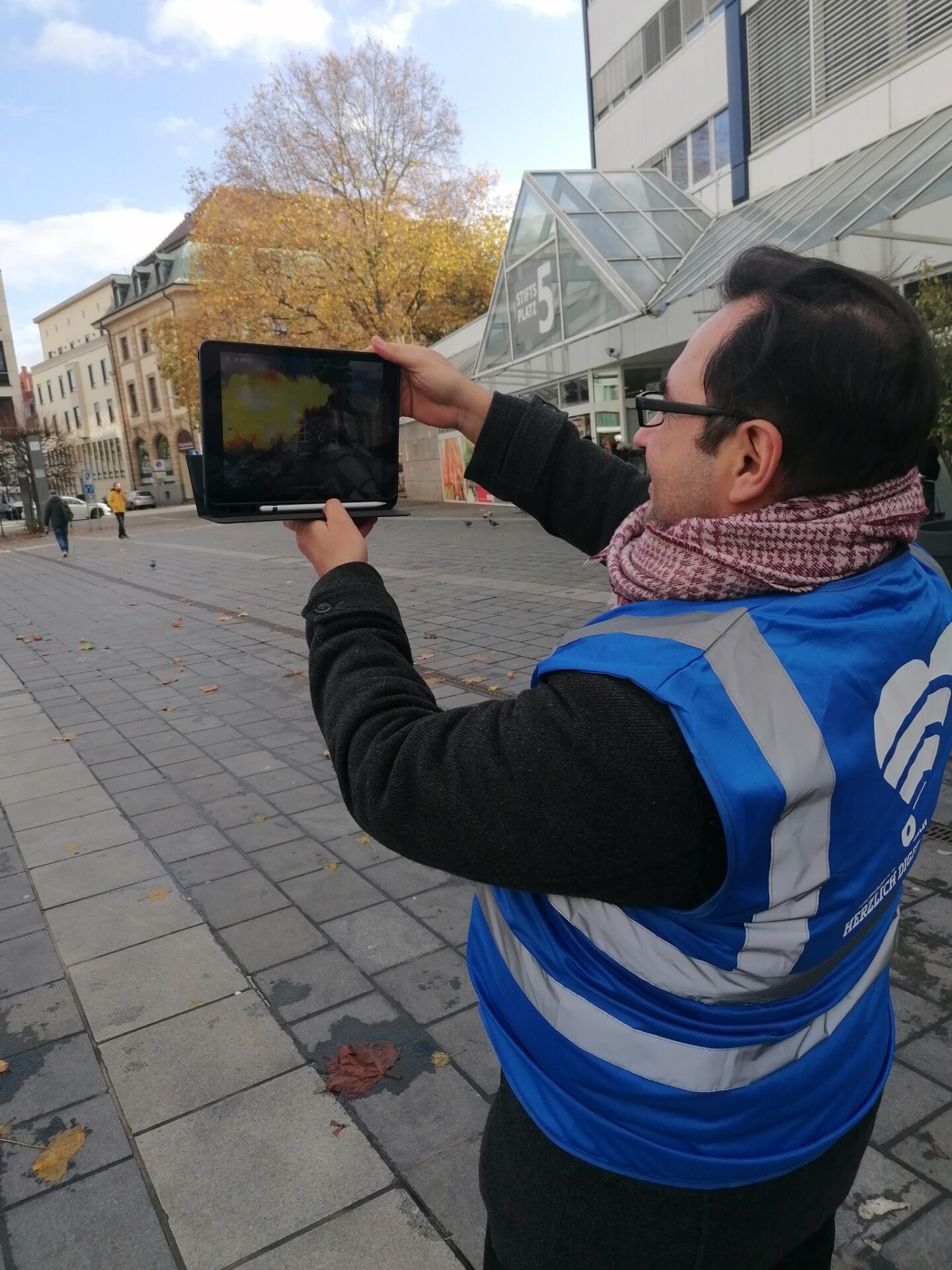 Ein Mann, der eine blaue Warnweste trägt, steht auf einem öffentlichen Platz und hält ein Tablet hoch. Auf dem Bildschirm sieht er einen feuerspeienden Drachen auf dem Platz.