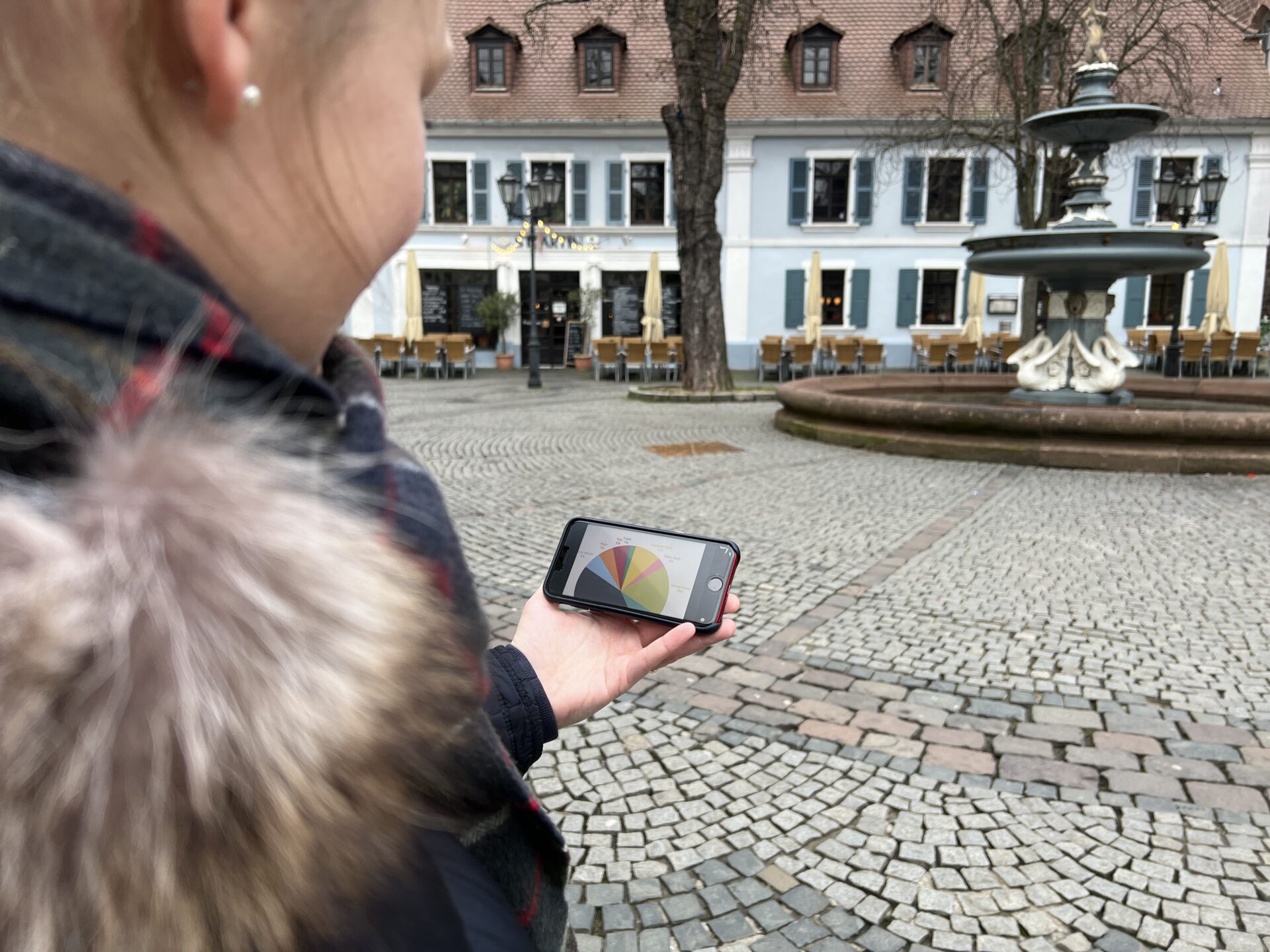 Eine Frau steht auf einem öffentlichen Platz vor einem Brunnen und schaut auf ein Handydisplay. Darauf ist ein Kuchendiagramm zu sehen.