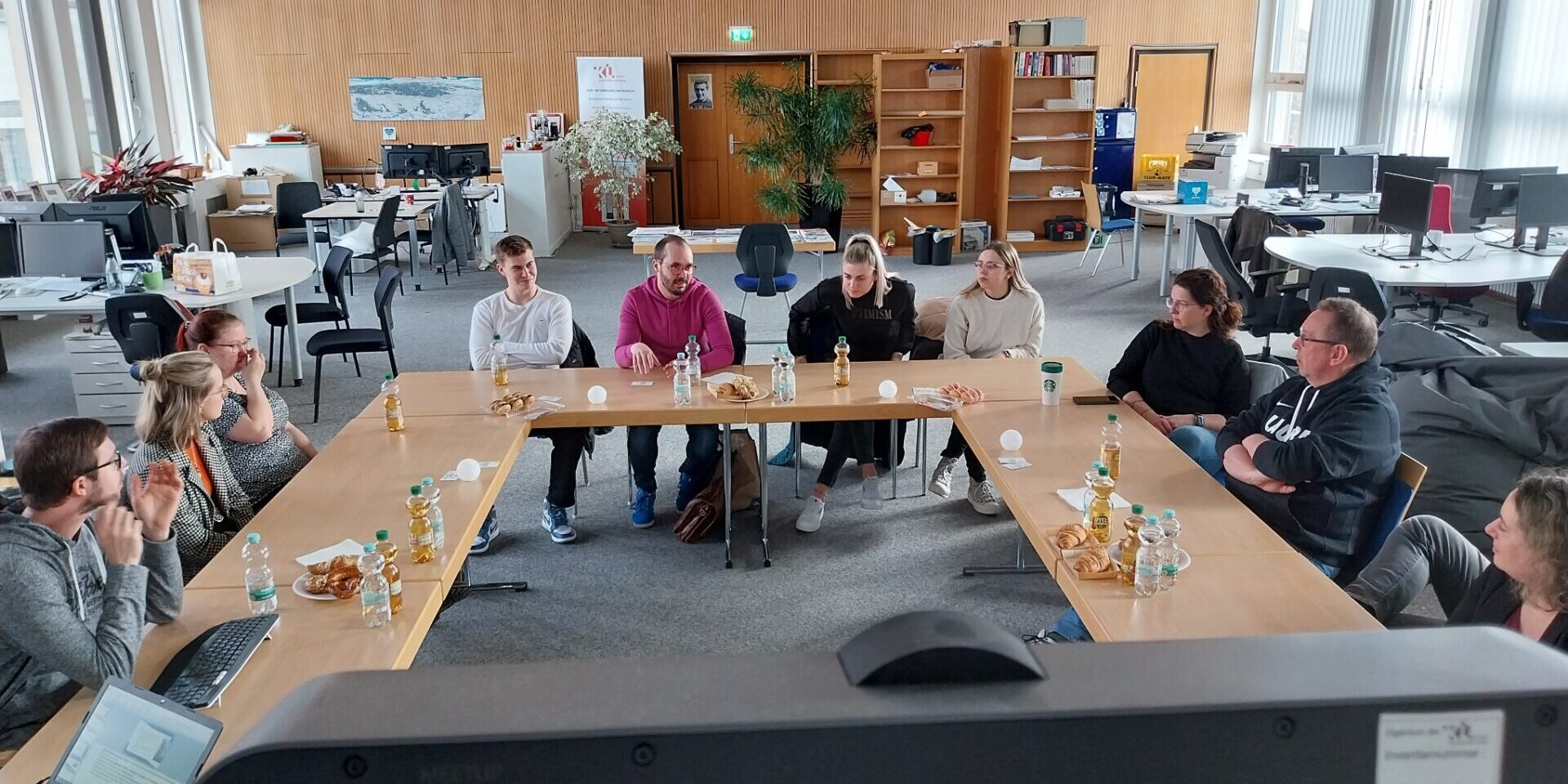 Zehn Menschen sitzen in einem Großraumbüro um eine u-förmige Aufstellung von Tischen. Es handelt sich um Mitarbeitende von Herzlich digital und Azubis der Stadtverwaltung.