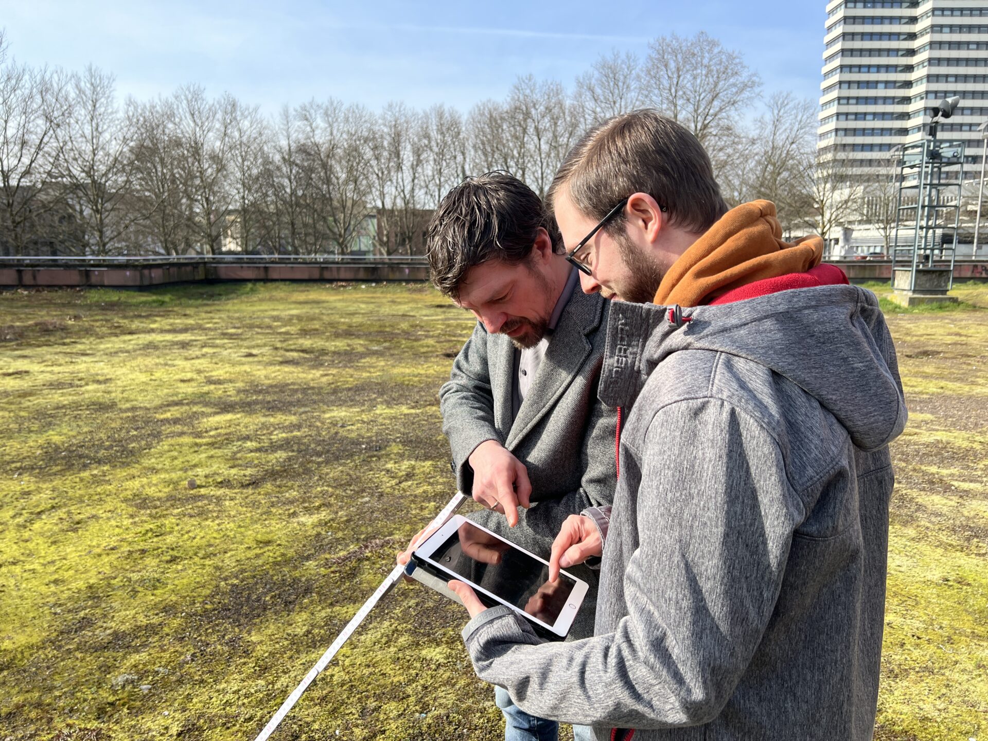 Zwei Männer stehen auf einer Wiese in der Stadt und betrachten etwas auf einem Tablet.