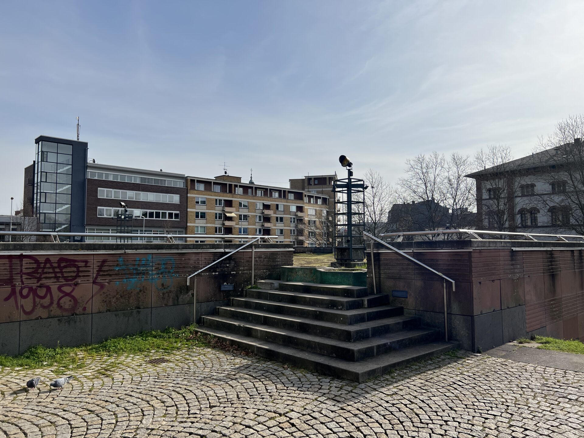 Ein Treppenaufgang zu einer Wiese, um die eine Mauer führt. Im Hintergrund größere Gerbäude einer Innenstadt.