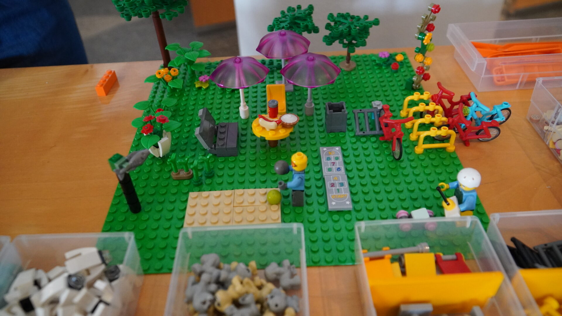 Aufbau aus Legosteinen zeigt einen öffentlichen Platz mit Schirmen, Sitzmöglichkeiten, Bäumen, Basketballfeld und Fahrrädern. Zwei Legofiguren spielen Basketball bzw. spielen mit einem ferngesteuerten Auto.