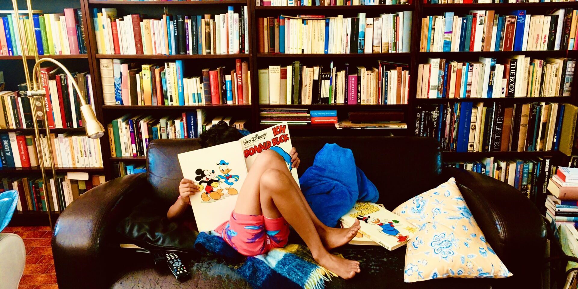 Ein Kind sitzt auf einem Sofa und liest ein Comicheft. Hinter dem Sofa steht ein Regal mit Büchern, das die gesamte Wand ausfüllt.