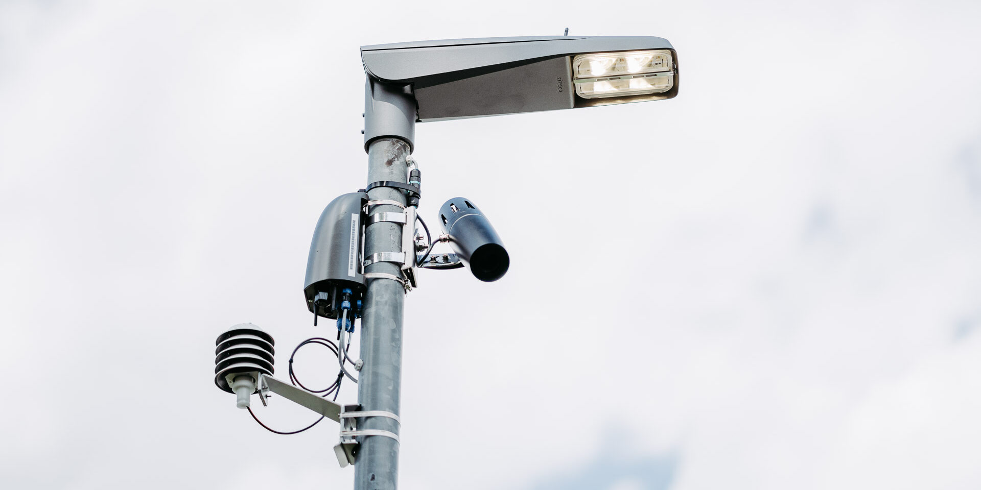 Eine leuchtende Straßenlaterne im Fauthweg vor grauem Himmel. Unterhalb des Laternenkopfes sind am Mast verschiedene Geräte angebracht.