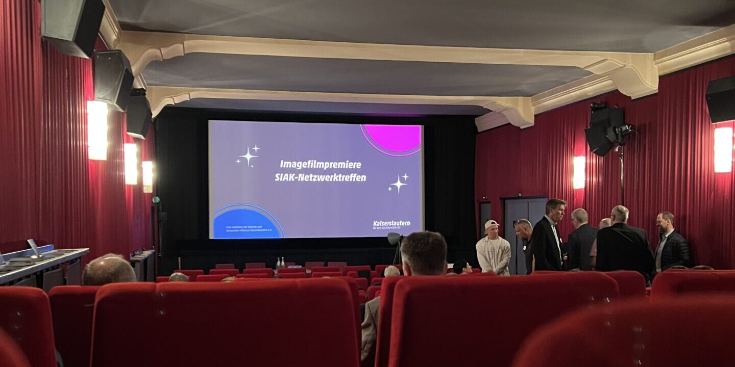 Ein Kinosaal mit roten Sesseln und Wänden. Auf der Leinwand steht "Imagefilmpremiere. SIAK Netzwerktreffen". Im Saal sind einige Plätze belegt, eine Gruppe Personen steht neben den Sitzreihen und ist im Gespräch.