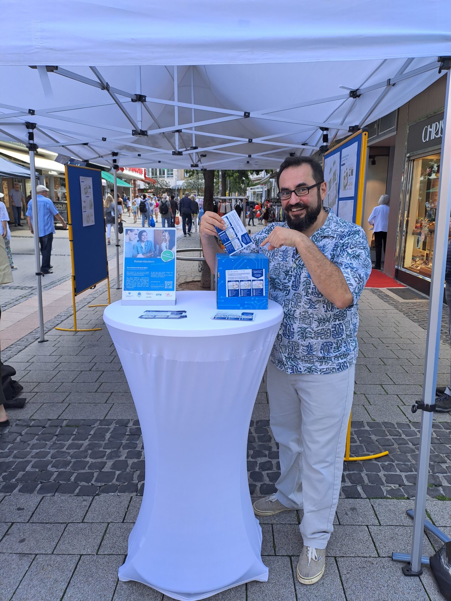 Ein Mann steht in einer Fußgängerzone in einem Pavillionzelt. Er wirft eine Postkarte in eine Box, die auf einem Stehtisch steht.