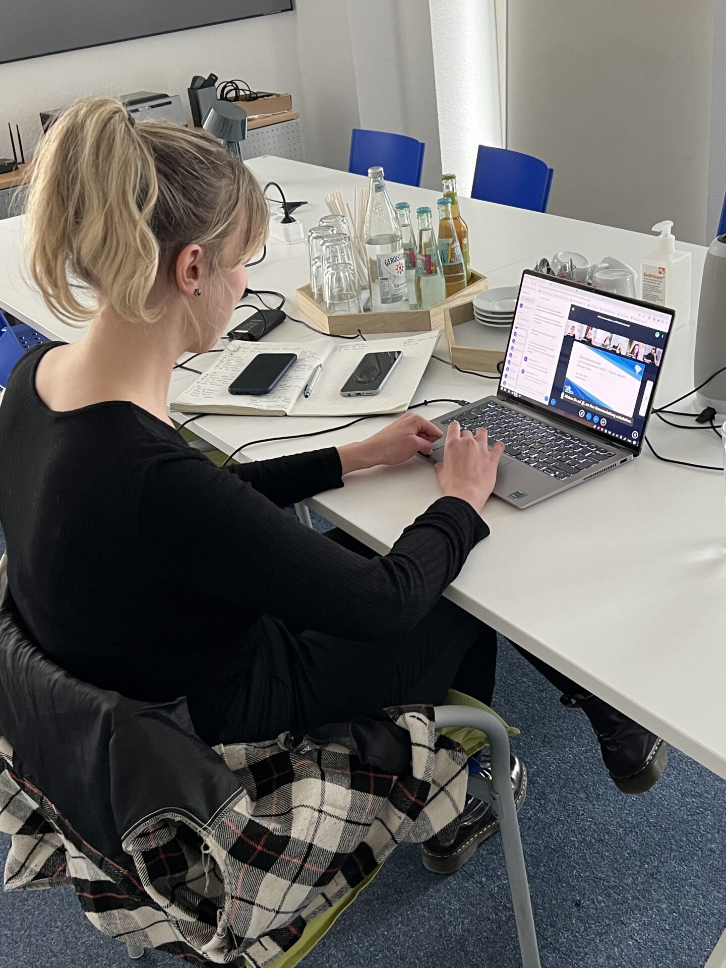 Eine Frau sitzt an einem Besprechungstisch und schaut auf ein Laptop, auf dem eine Videokonferenz läuft.