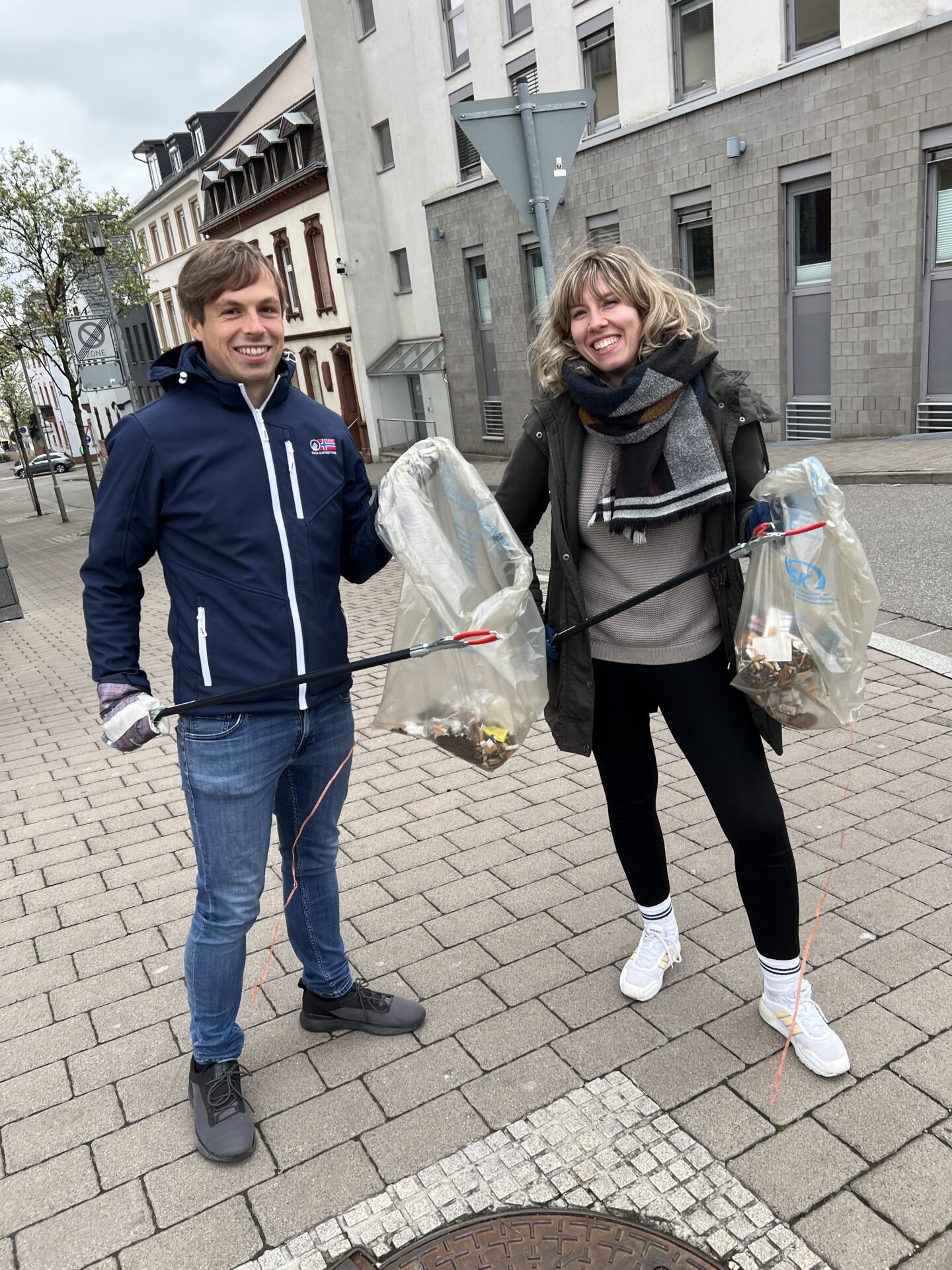 Ein Mann und eine Frau posieren für ein Foto. Sie haben Müllbeutel und Müllzangen in der Hand.