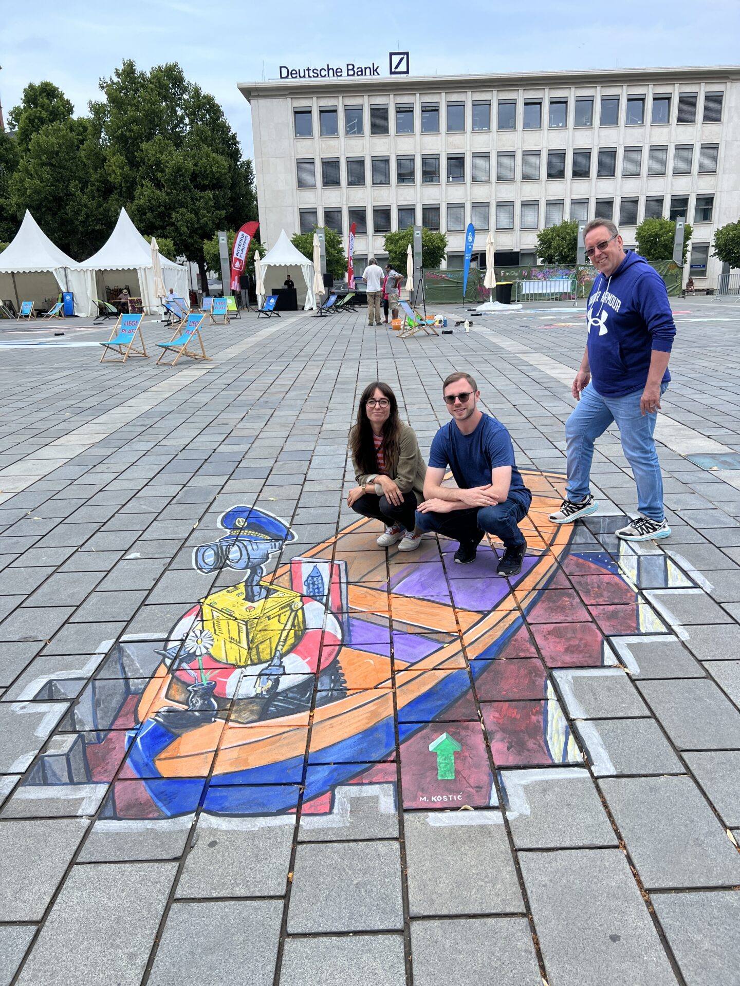Eine Frau und zwei Männer posieren auf einem Straßenkunstwerk auf einem öffentlichen Platz. Das Kunstwerk zeigt ein Boot, das von einer Art Roboter gesteuert wird.