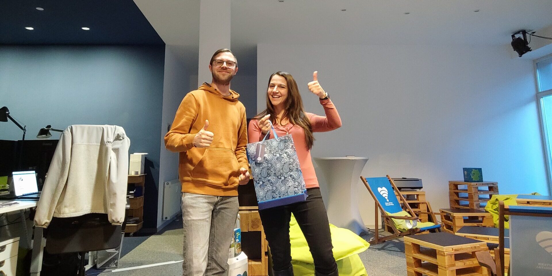 Ein Mann und eine Frau stehen in einem Raum. Sie hat beim Gewinnspiel der Beteiligungsplattform gewonnen und posiert mit einer Geschenktüte.